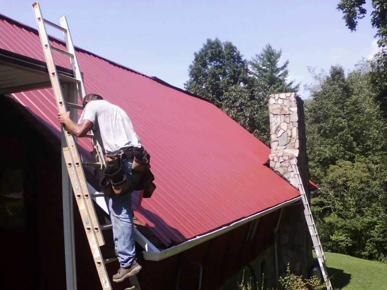 Roofer on a ladder beside a red metal roof and stone chimney — Soffit & Fascia Repair by Roofing Brothers in Glenns Ferry